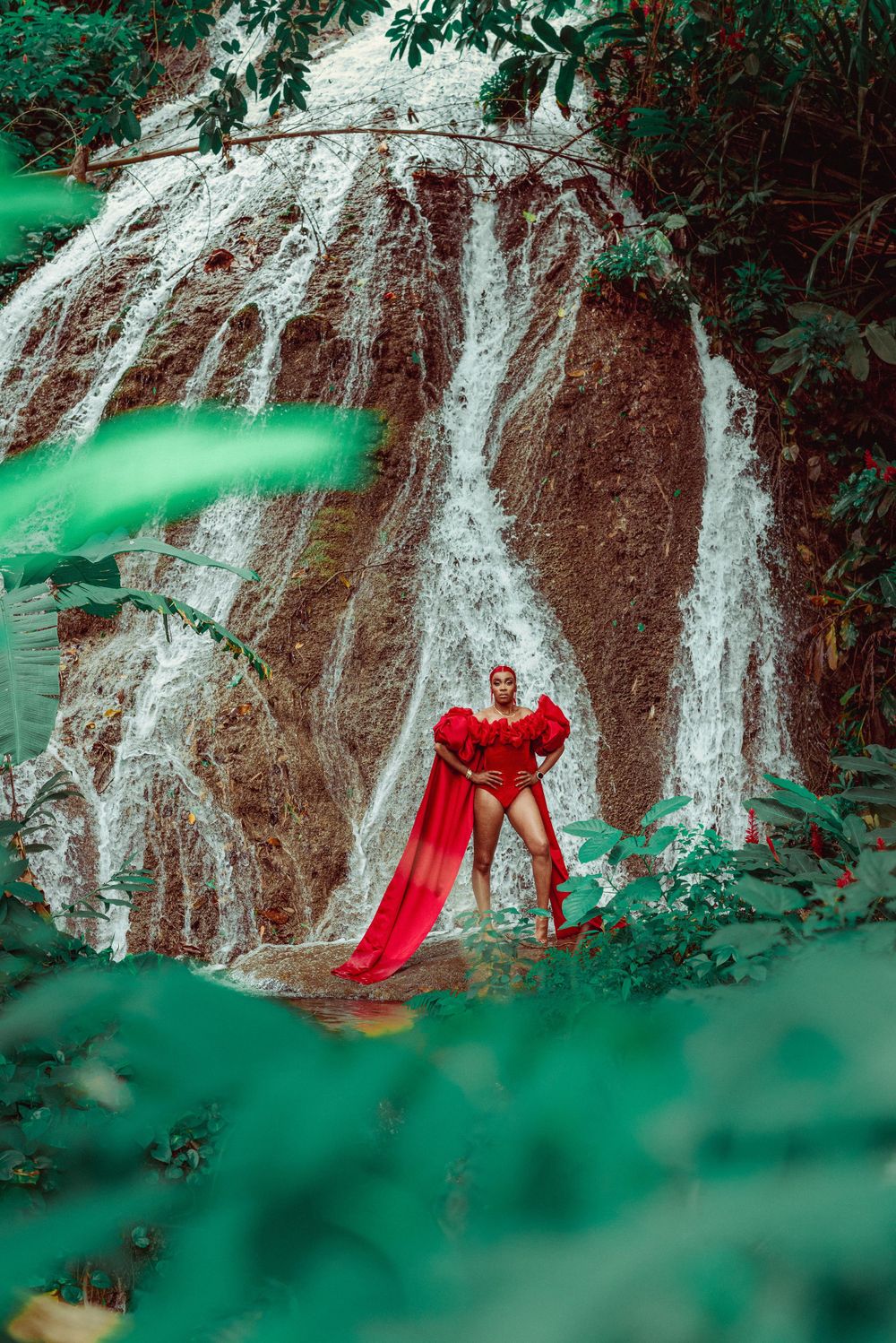 Dramatic portrait in red flowing dress against a cascading waterfall with vibrant green foliage.