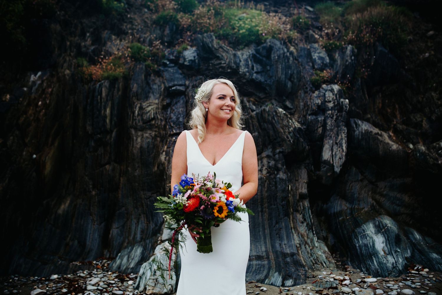 A bride in white dress holds a colorful bouquet against dramatic black rock cliff backdrop.