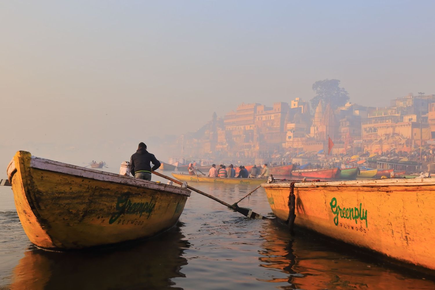 Wooden boats float on the misty Ganges River with ancient buildings rising in the background.
