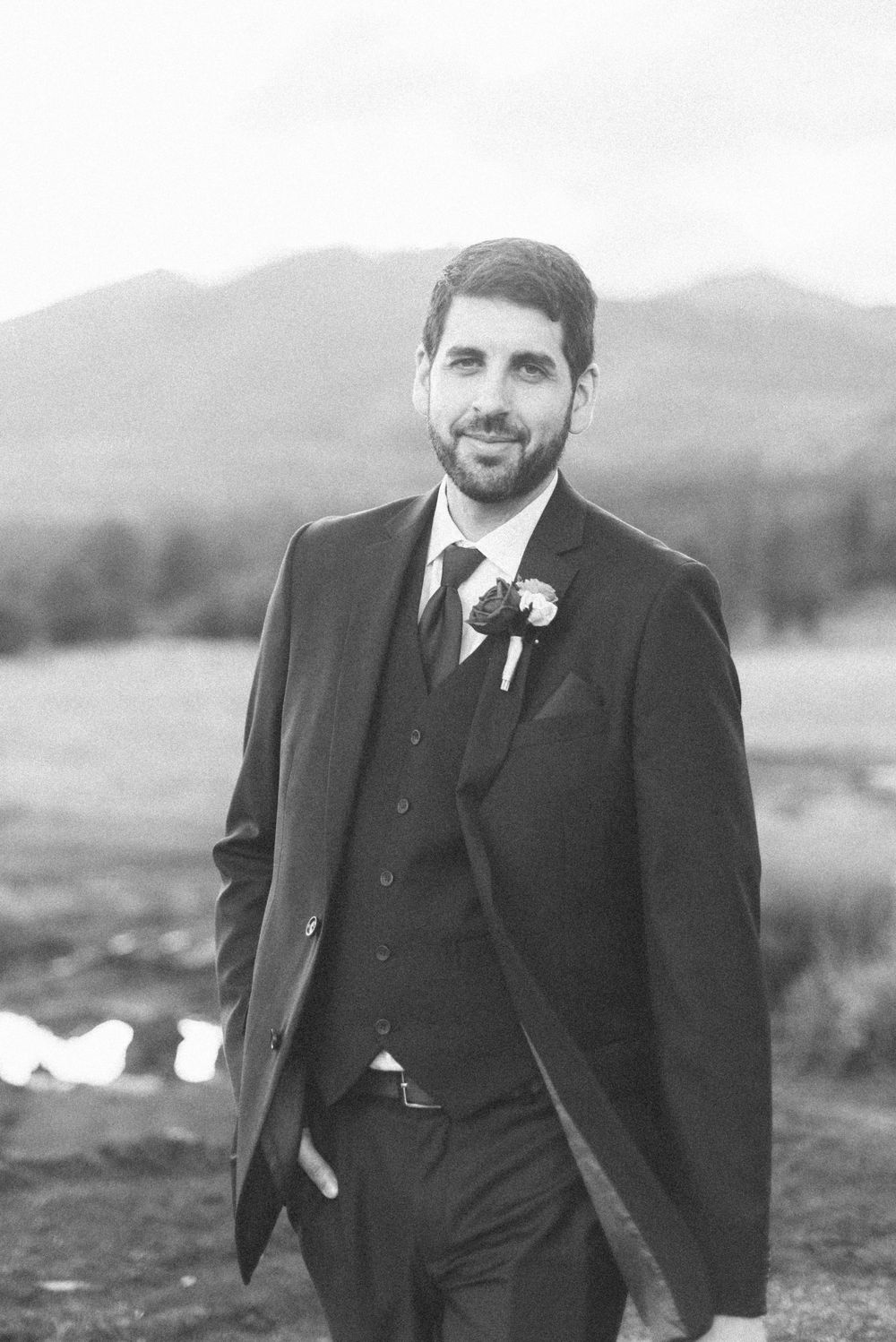 Black and white portrait of groom in formal suit with mountain landscape background.