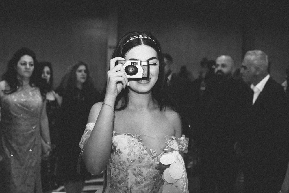 Black and white photo of bride taking picture with vintage camera during reception.