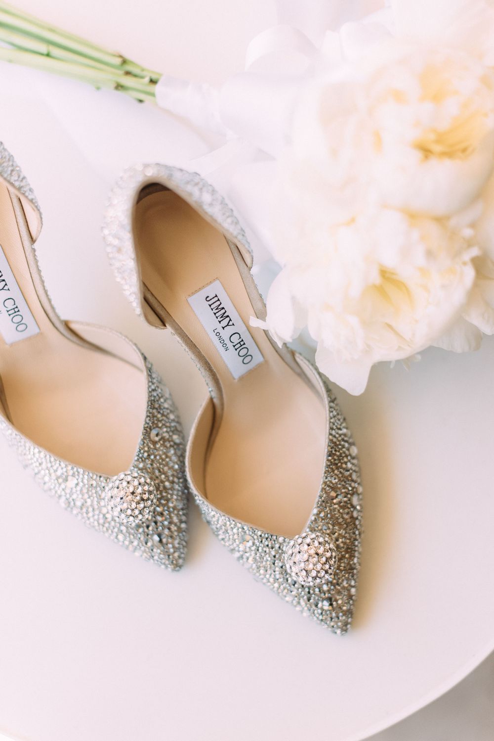 Sparkly silver Jimmy Choo bridal shoes next to light pink flowers on white surface.