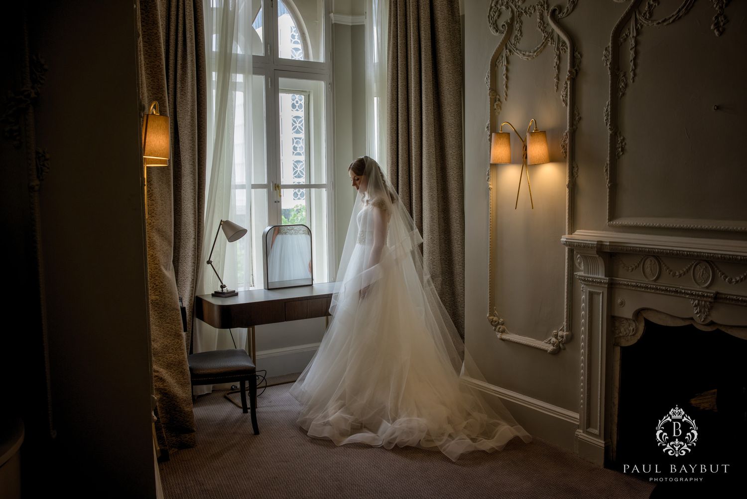 Elegant wedding dress silhouette against arched window in vintage room with wall sconces.