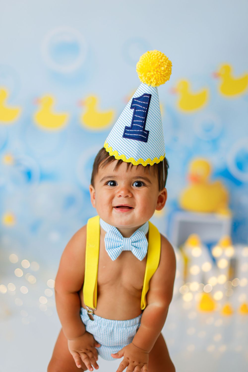 First birthday party setup with yellow pom pom hat, blue bow tie and yellow suspenders against rubber duck theme backdrop.
