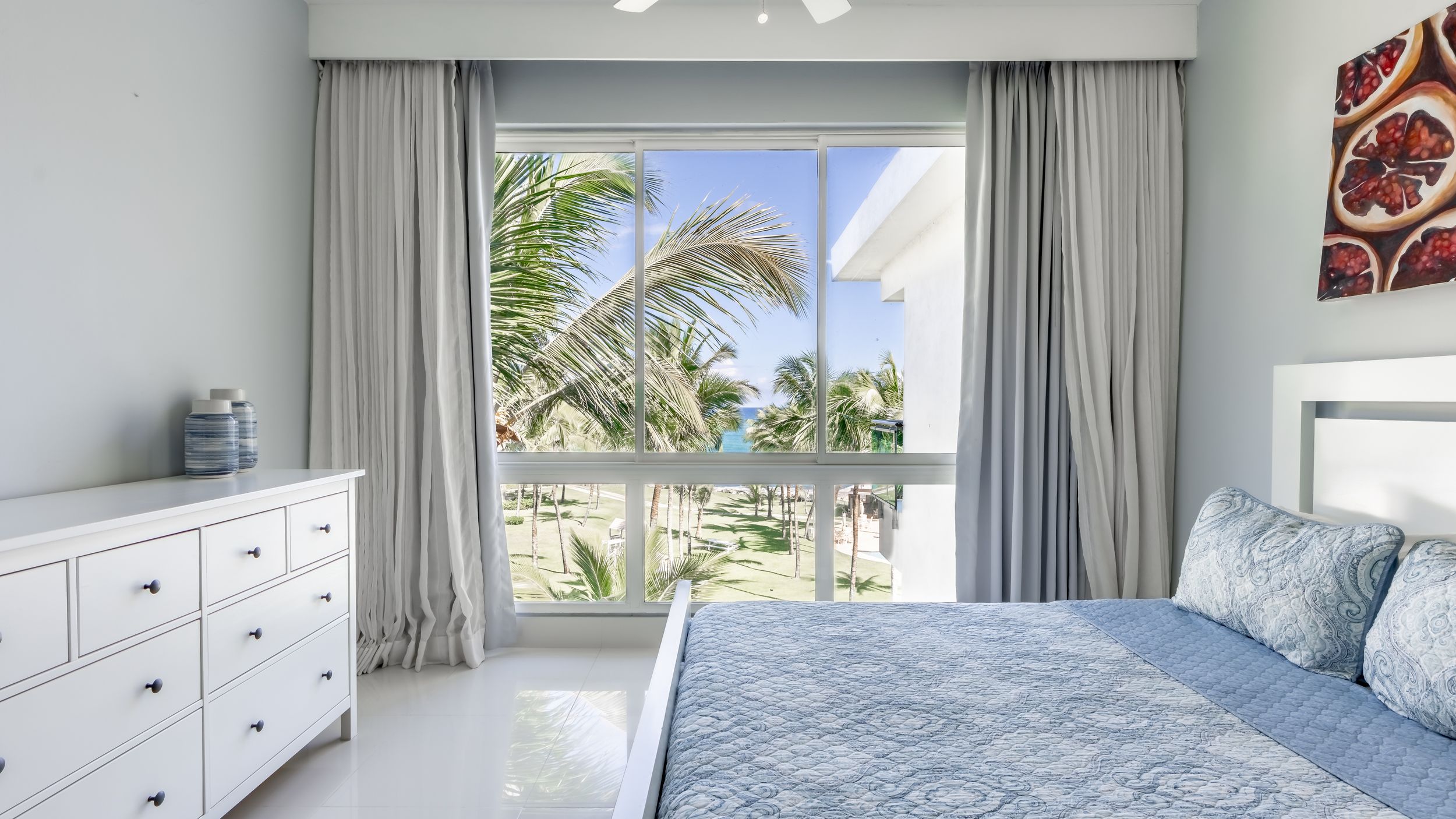 Modern beachfront bedroom with white furniture and palm tree views through sliding glass doors.