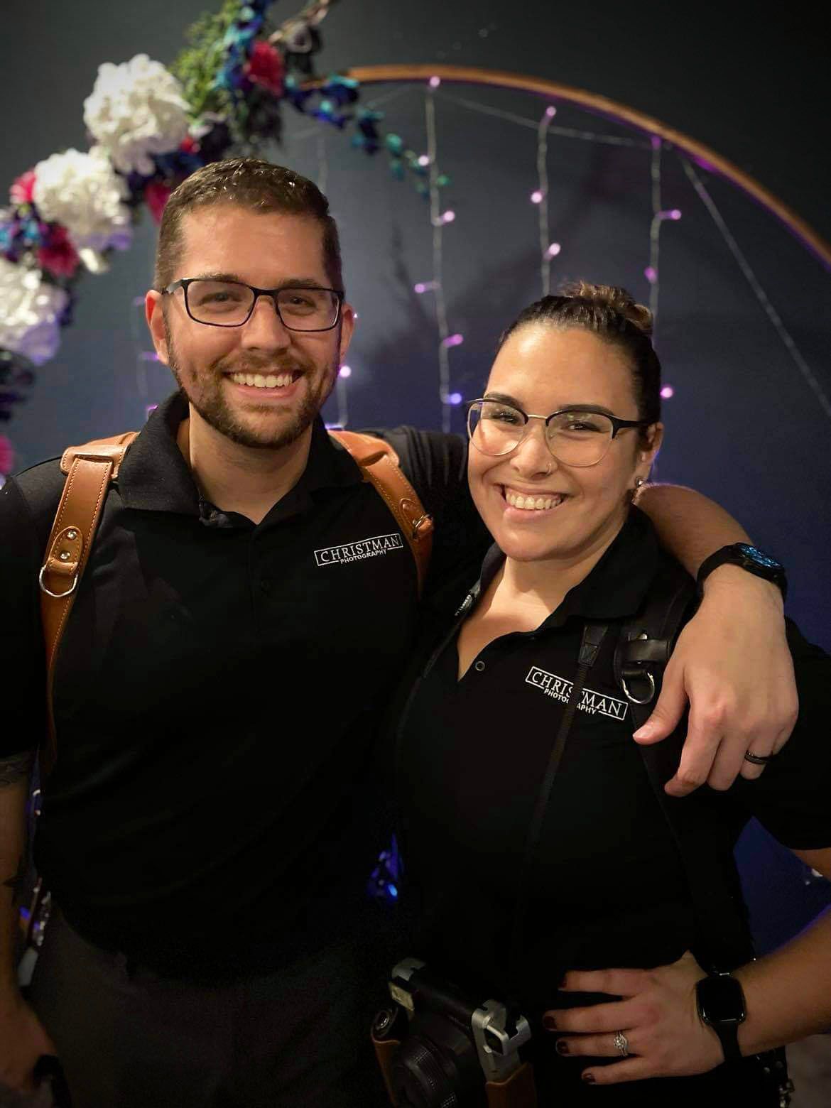 Two photographers in matching black uniforms pose together against a floral backdrop.