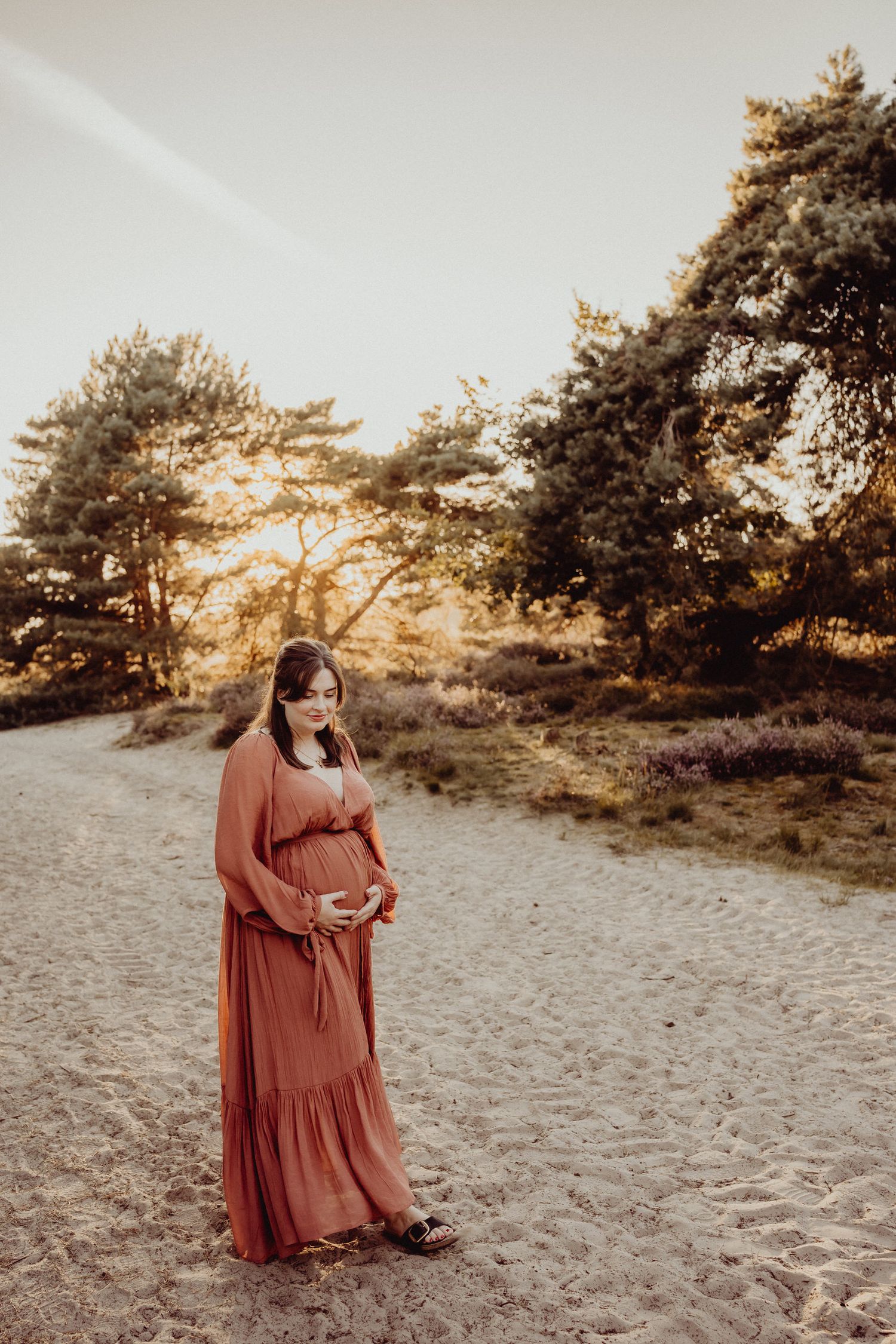 A rust-colored maxi dress photographed in a natural outdoor setting with pine trees at sunset.
