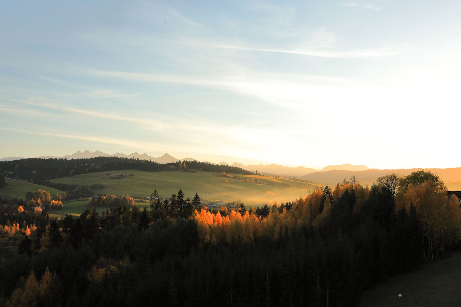 Misty mountain landscape at sunset with golden forest and rolling hills in the distance.