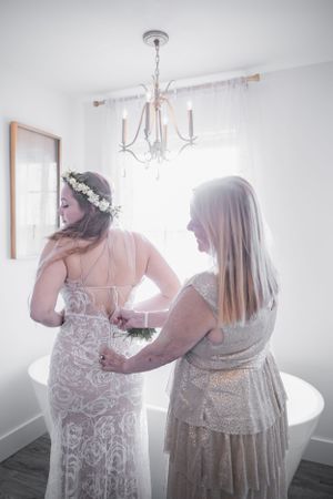Two people help with the intricate lace details of a wedding dress in a sunlit room.