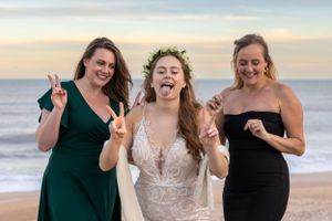 Three people in formal attire celebrate joyfully on a beach at sunset.