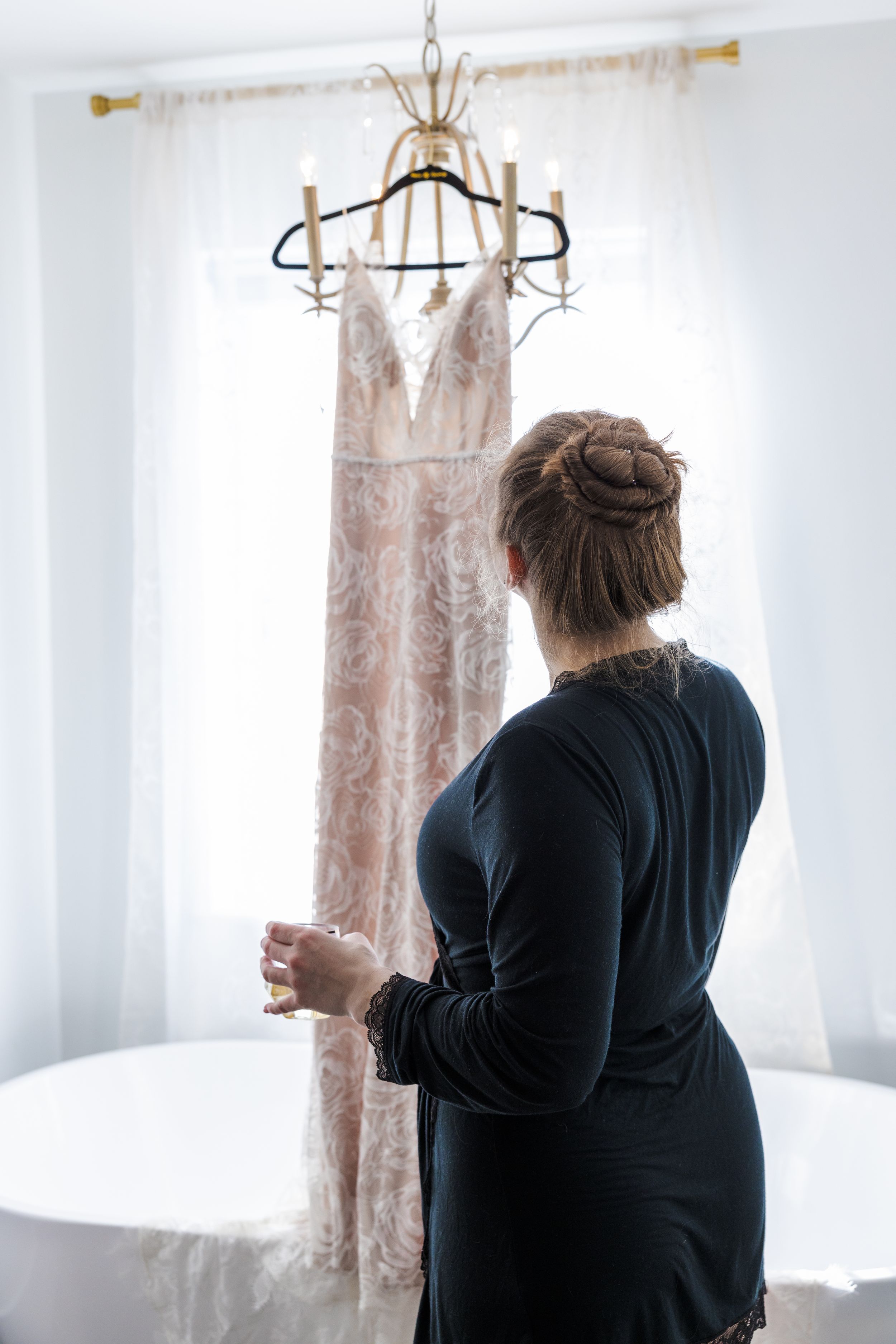 A delicate lace wedding dress hangs from an elegant chandelier as someone in black admires it.