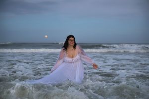 A figure in a white dress stands in ocean waves at sunset with moody stormy skies in the background.