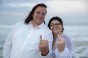 A couple shows off their rings while posing playfully by the ocean.