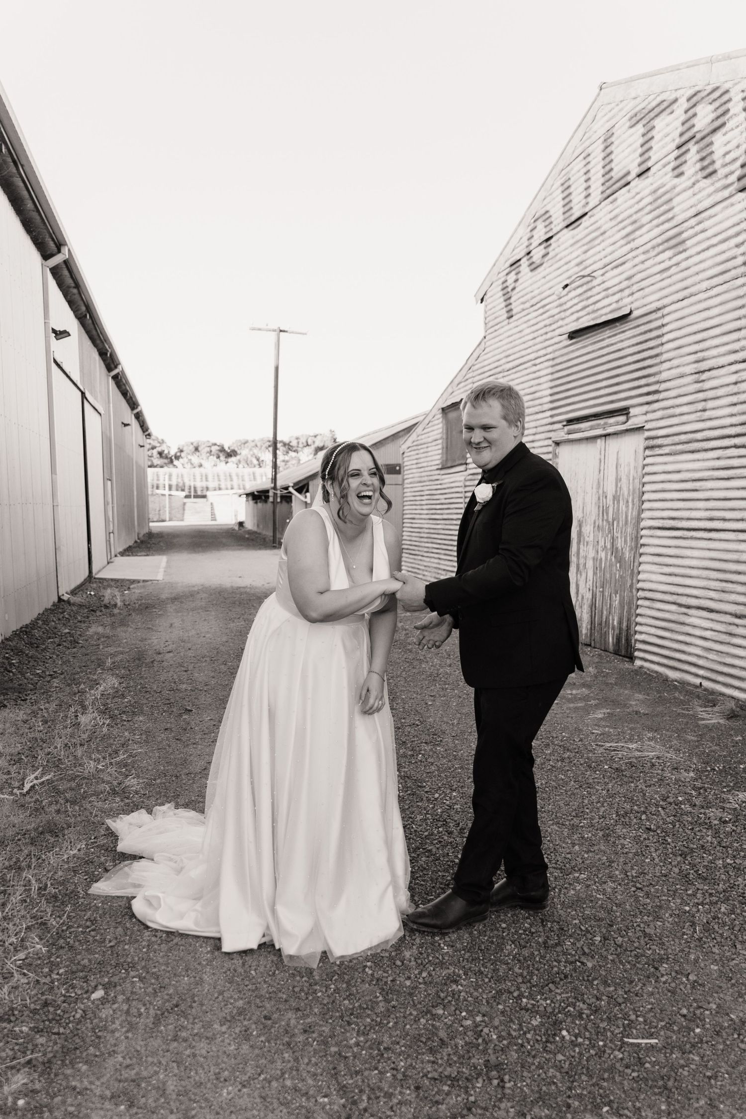 A joyful wedding couple shares a laugh outside a rustic industrial building in black and white.