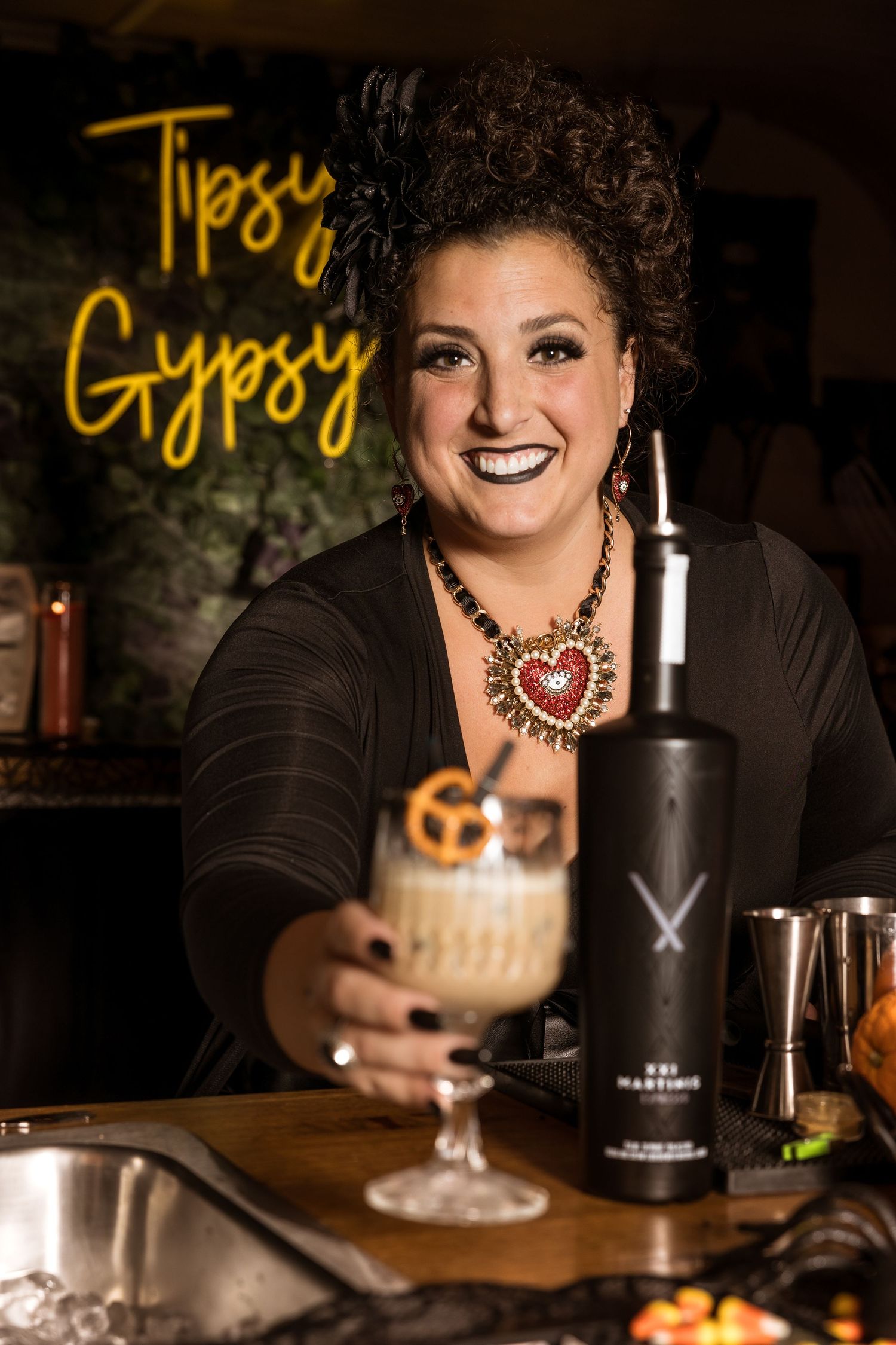 A bartender serves specialty drinks under a neon Tipsy Gypsy sign at a Halloween-themed event.