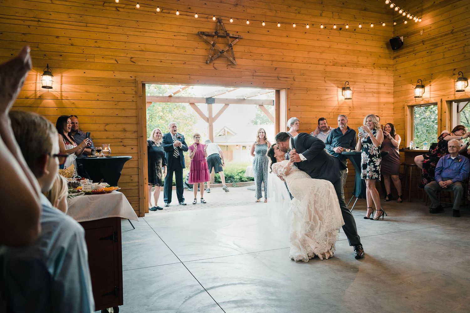 Wedding dance captured in a wooden barn with string lights and natural light streaming through doorway.