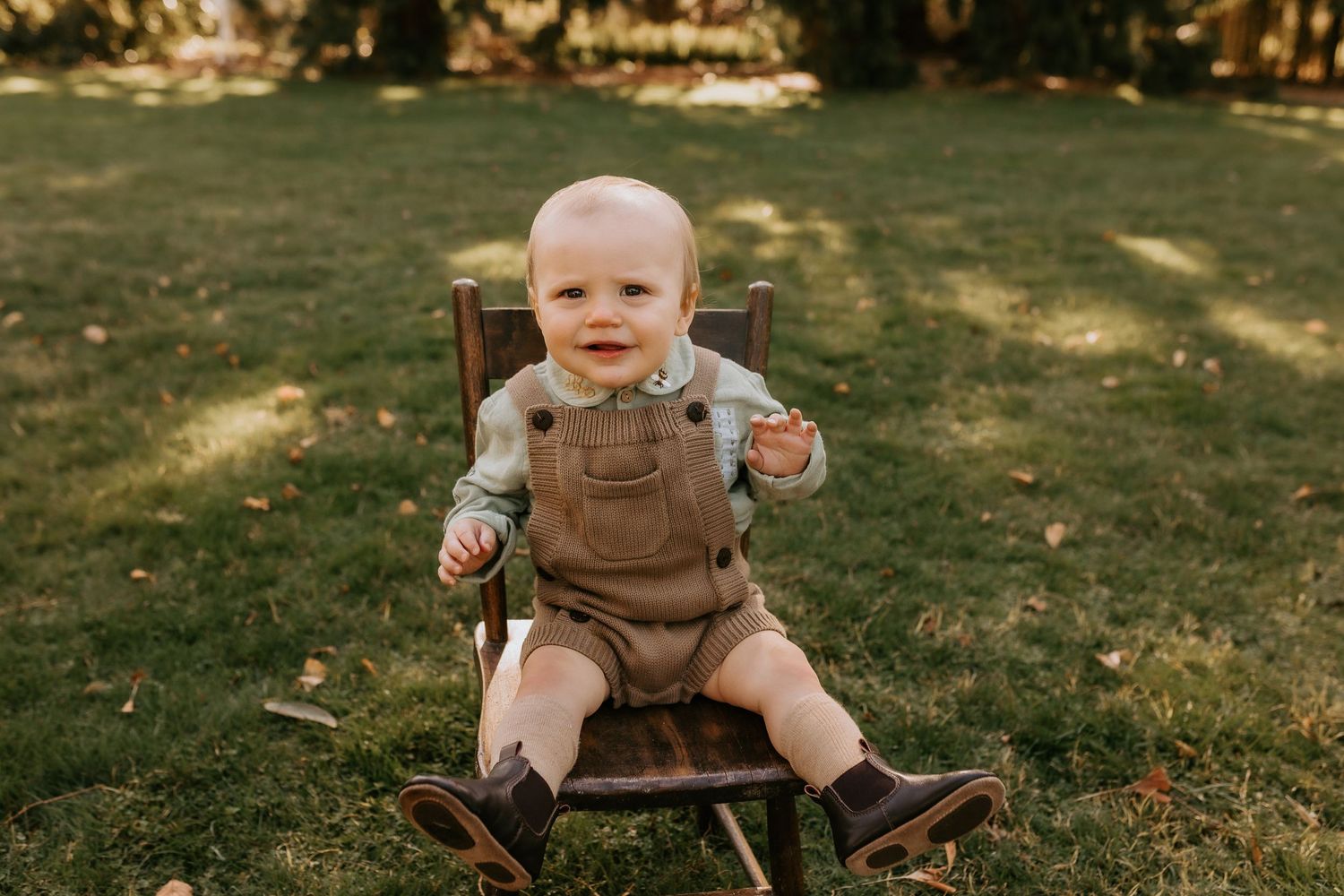 Baby wearing brown overalls sits on grass during outdoor autumn photoshoot.