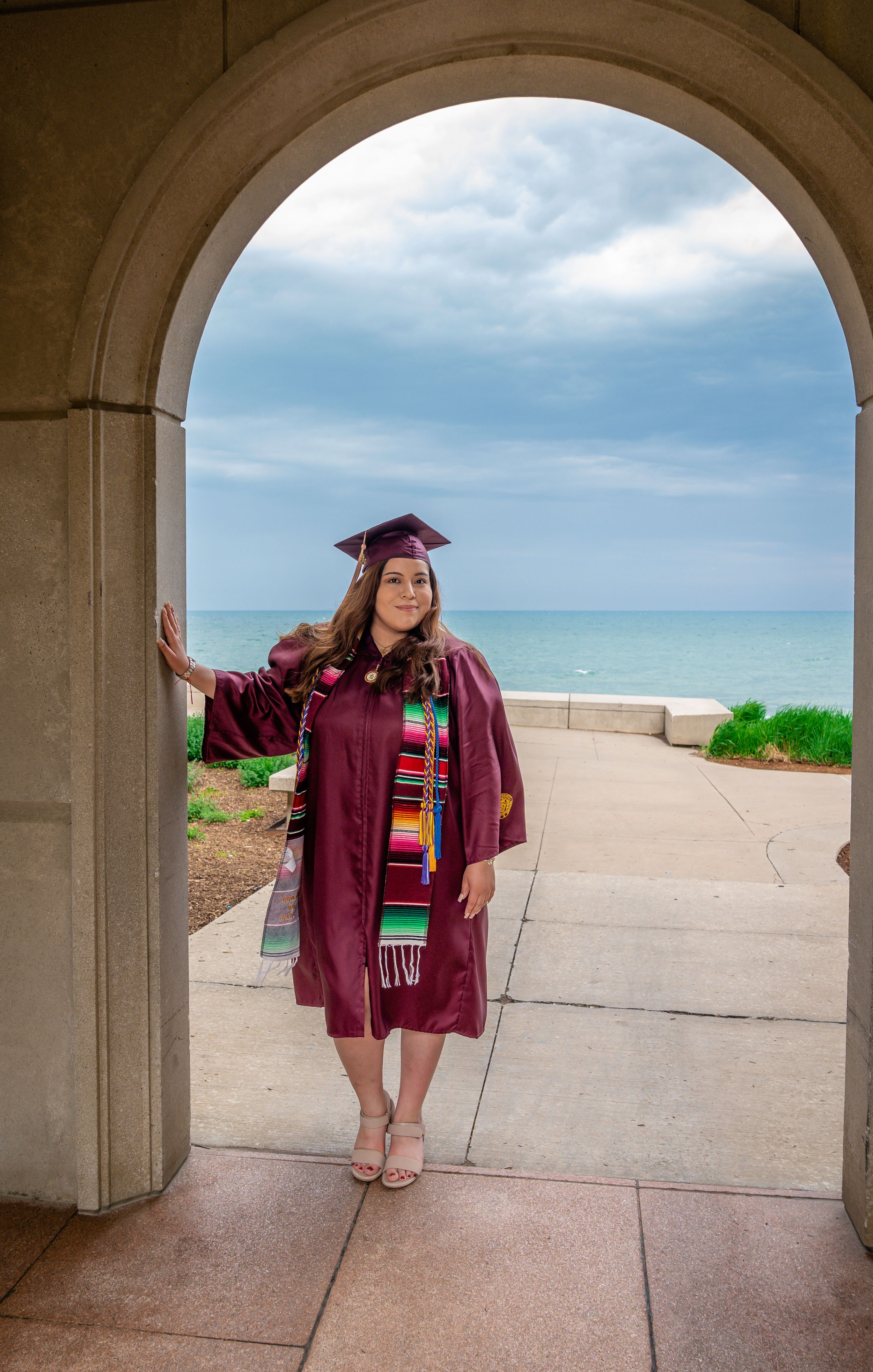 Graduate in maroon regalia poses under an arched walkway with beach and ocean views.