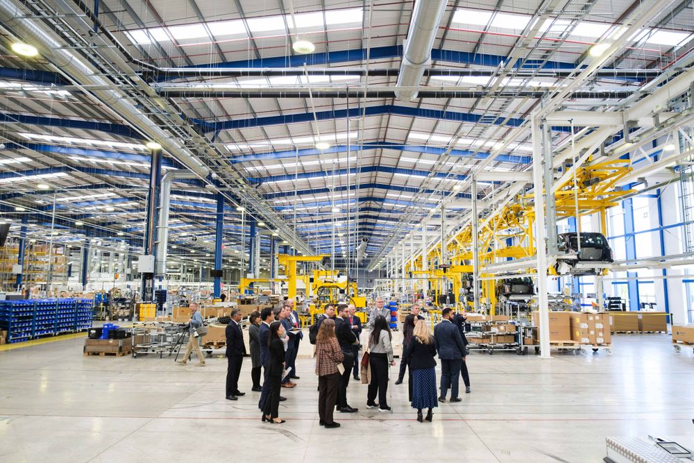 Group of visitors touring a modern industrial factory floor with yellow robotic equipment and high ceiling.
