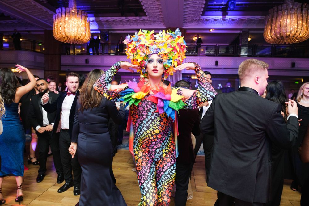 Colorfully costumed performer dancing among formal party attendees in ballroom setting.