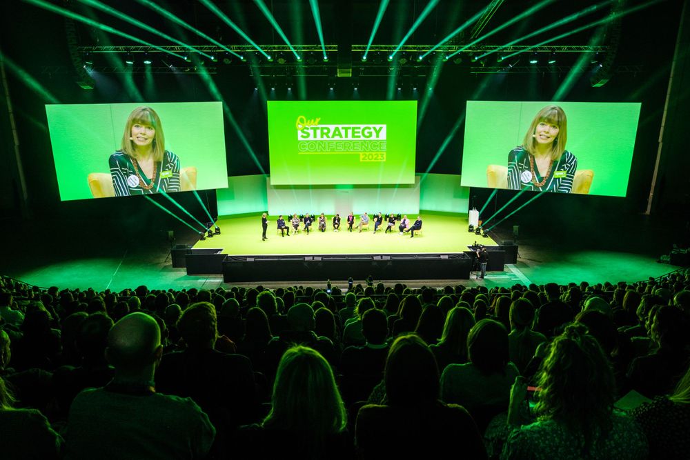 Large conference auditorium with green stage lighting and presentation screens displaying keynote session.