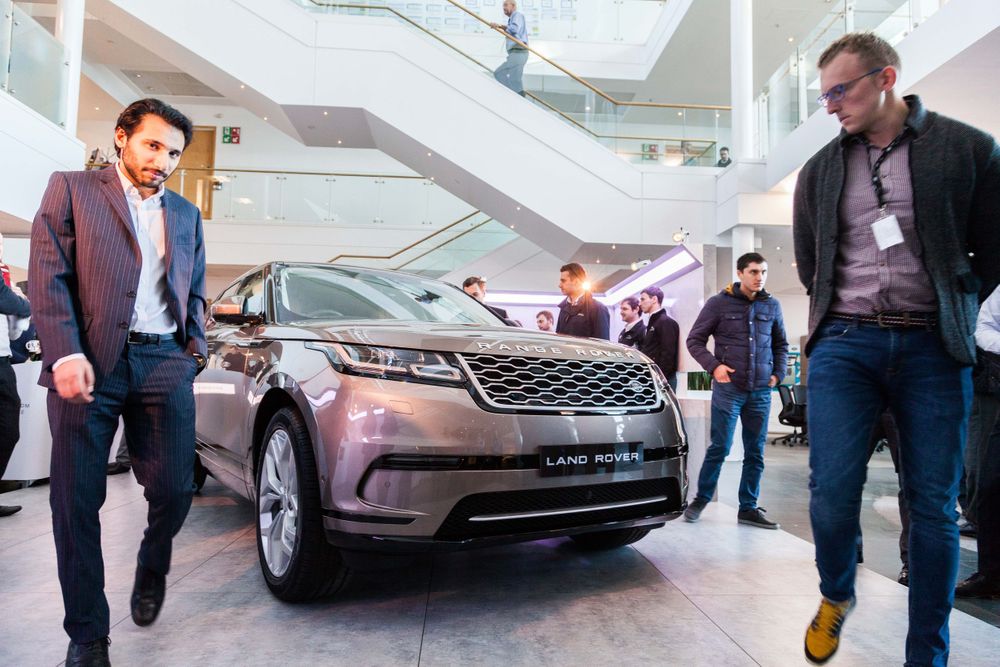 Group examines new silver Range Rover vehicle in modern showroom with escalators.