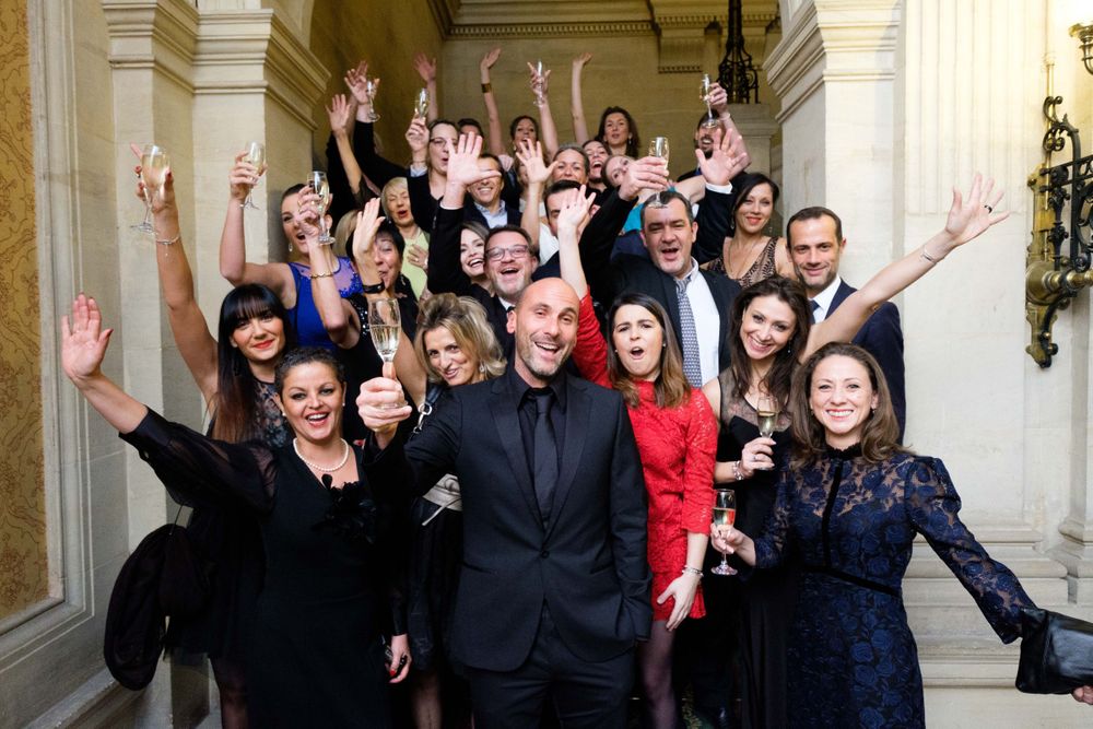 Large group poses together in formal attire with raised arms in elegant hallway setting.