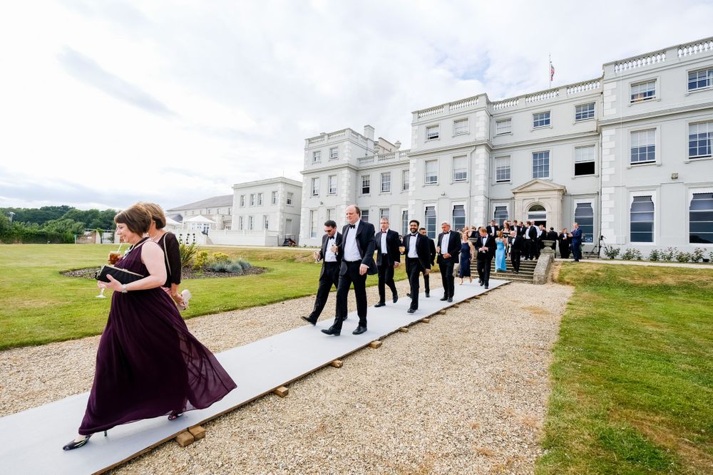Wedding party walking along path outside elegant white mansion on sunny day.