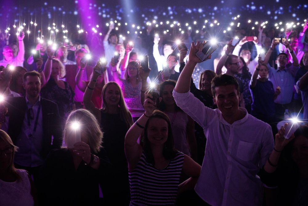 Concert crowd holds up glowing phones creating sea of lights during nighttime performance.