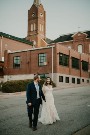 A wedding couple walks together near a historic brick church with clock tower at dusk.