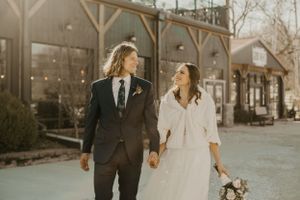 A couple walking together in wedding attire along a rustic building exterior.
