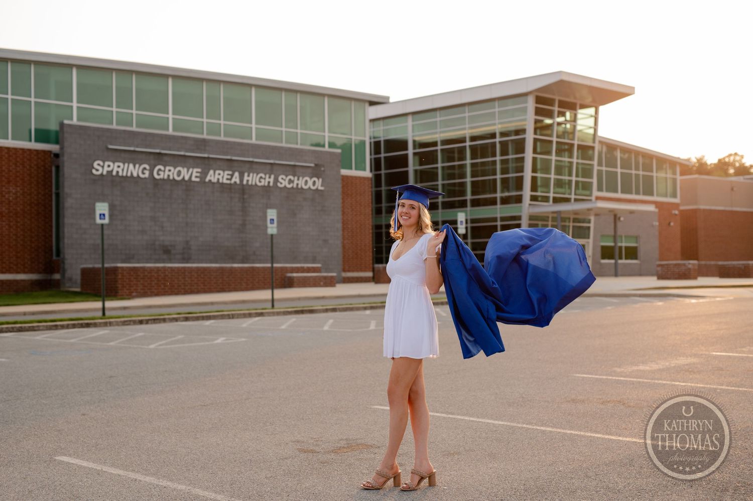 Graduate in white dress and blue gown posing outside high school at sunset.