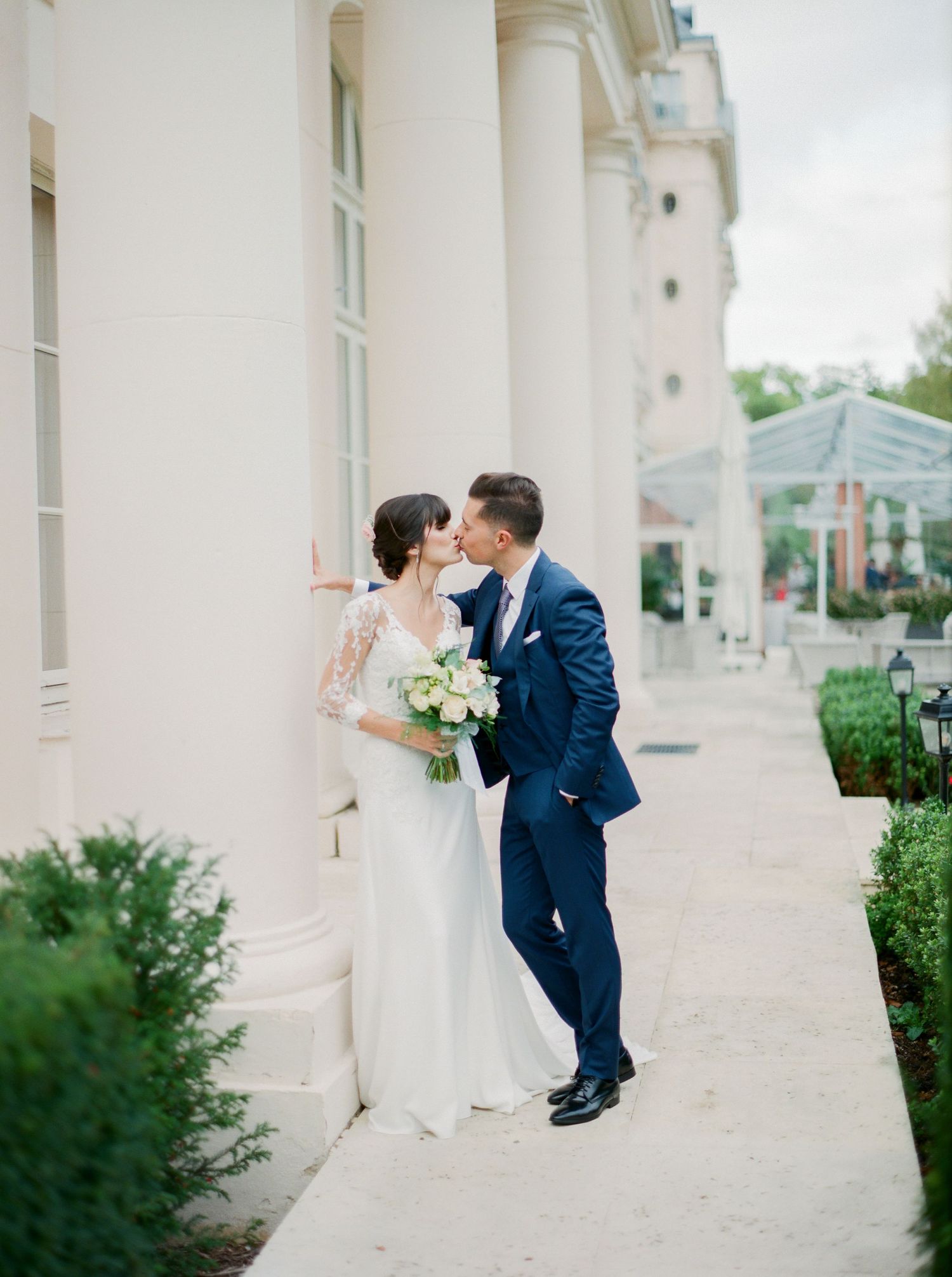 Une mariée en robe blanche tient un bouquet tout en partageant un moment romantique avec son marié près de colonnes blanches élégantes.