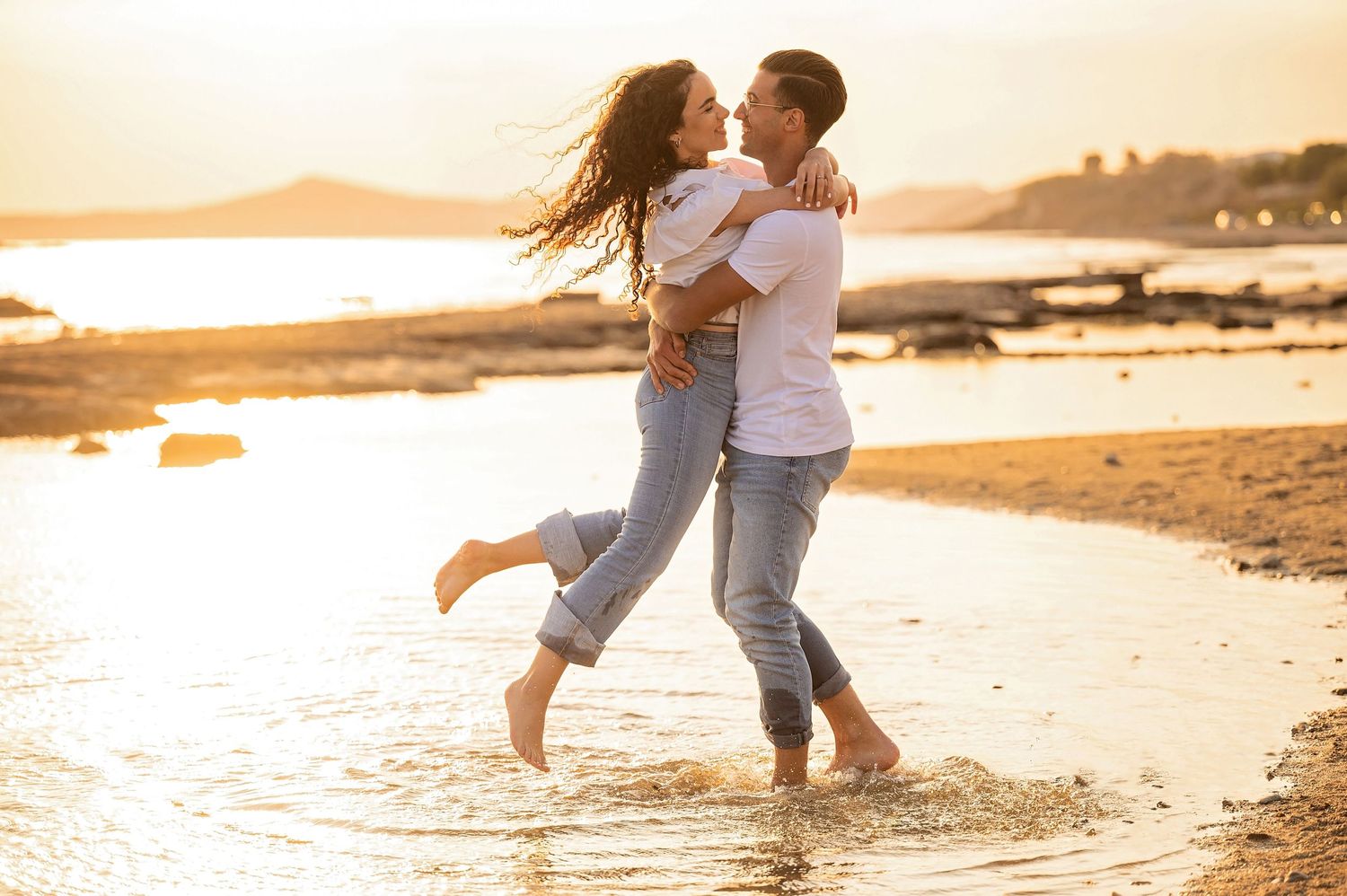 A romantic couple dances and embraces in shallow ocean water during golden hour at the beach.