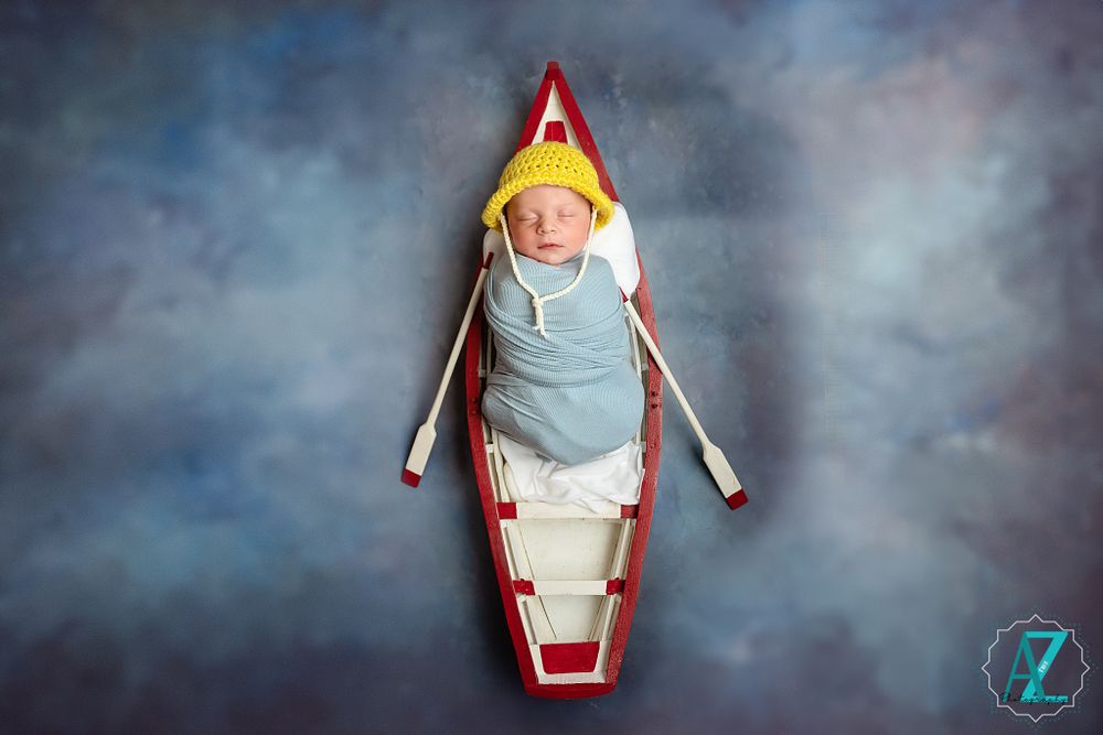 Newborn baby sleeping in a red and white canoe wearing a yellow hat against misty backdrop.
