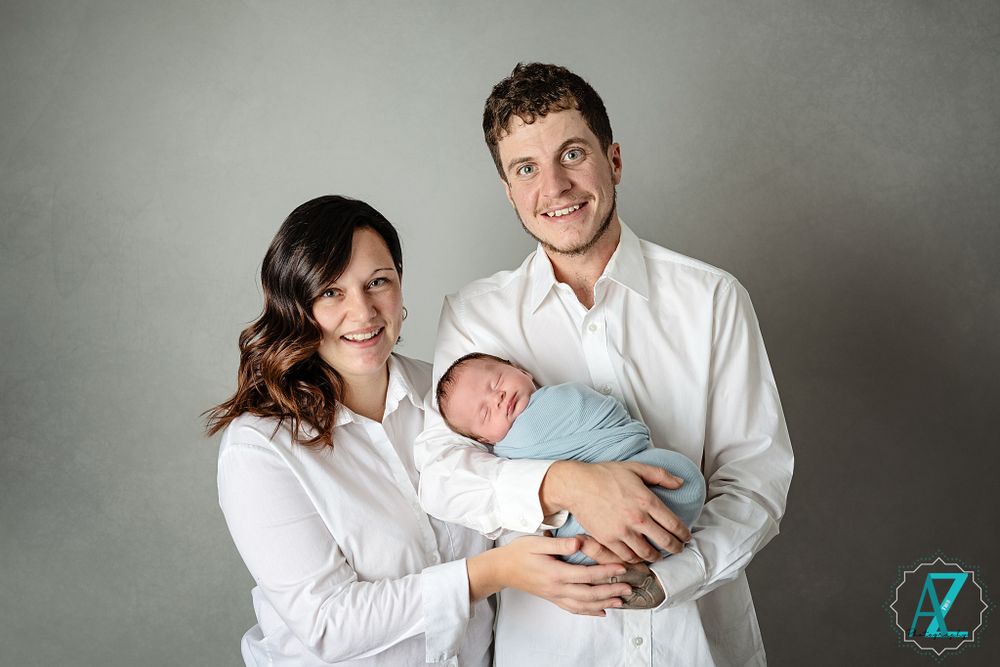 A couple in white shirts hold their newborn baby wrapped in light blue against a gray studio backdrop.
