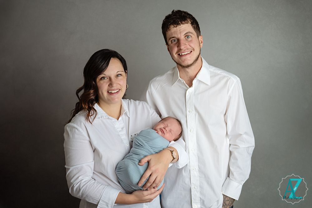 Family portrait featuring parents in white shirts holding their sleeping newborn against gray background.