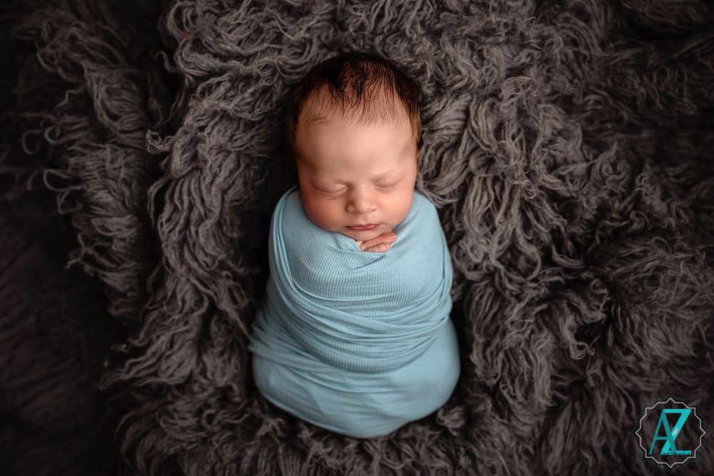 Overhead view of sleeping newborn swaddled in light blue on dark textured backdrop.