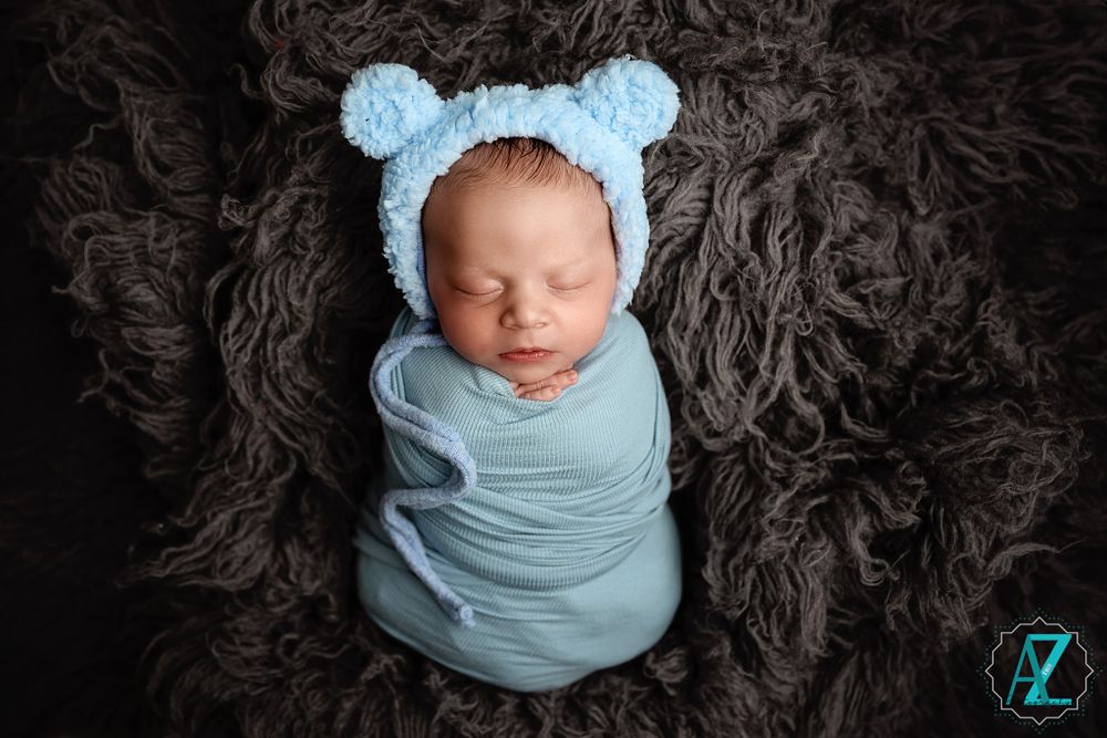 Sleeping newborn wrapped in blue wearing a knit bear hat on dark textured blanket.