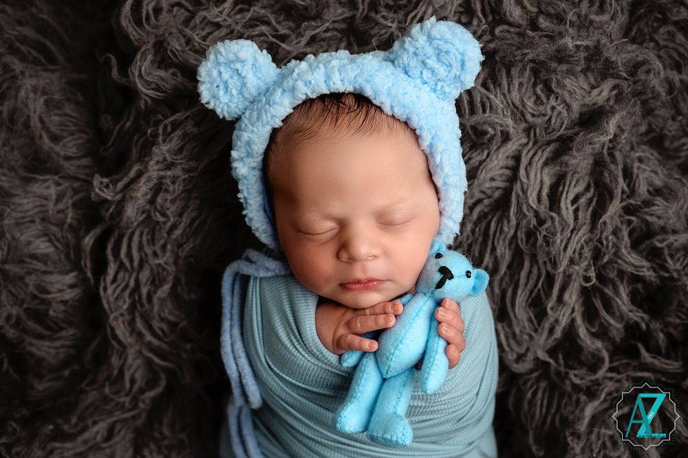 Sleeping newborn in blue swaddle and bear hat holding small plush toy on dark fabric.