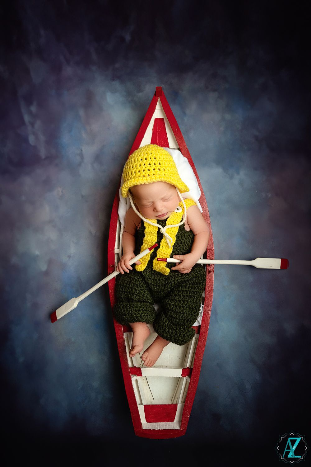 Newborn in yellow hat and green outfit sleeping in red and white canoe with paddle.