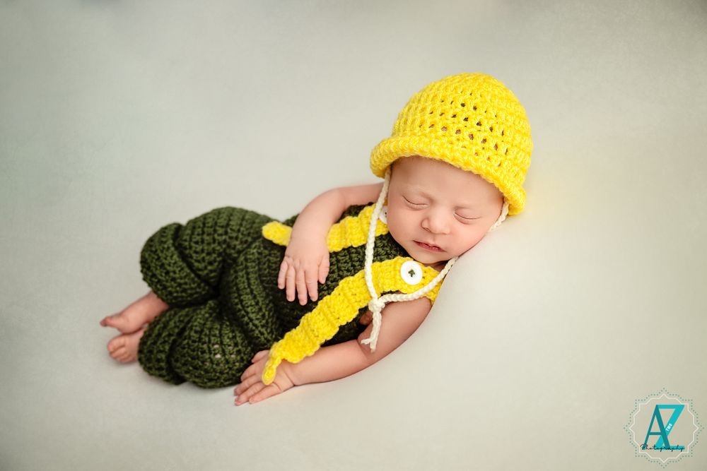Sleeping newborn in yellow hat and green overalls lying peacefully on light backdrop.