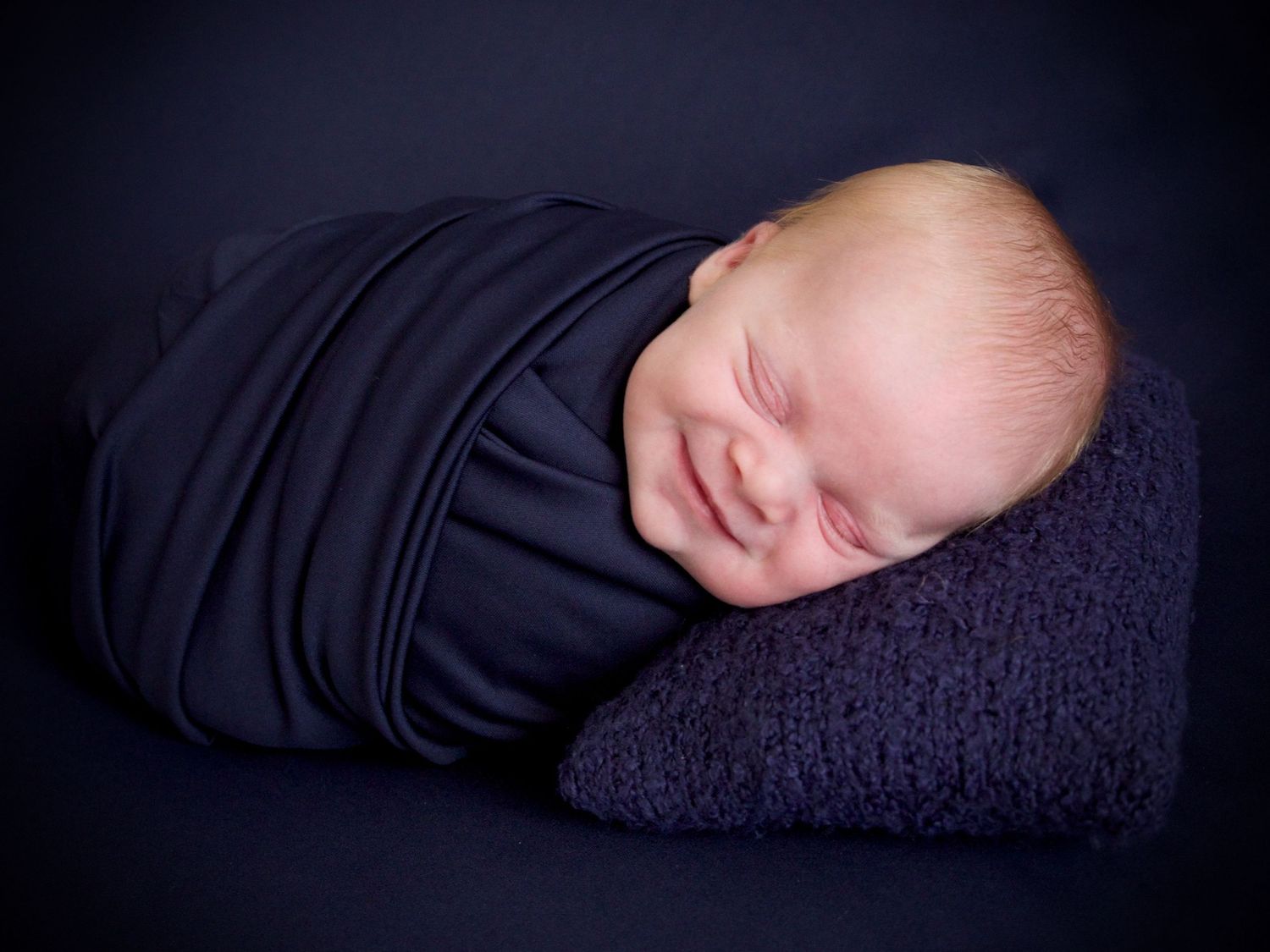 portrait of newborn baby boy in blue with big smile on his face laying on a pillow