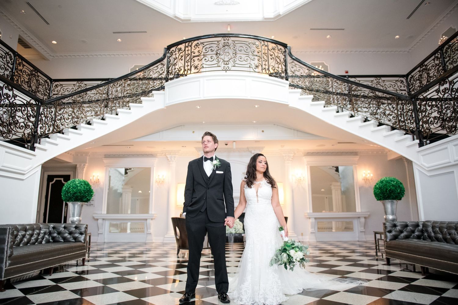Bride and groom holding hands in an elegant ballroom with ornate staircase and black and white tile floor.
