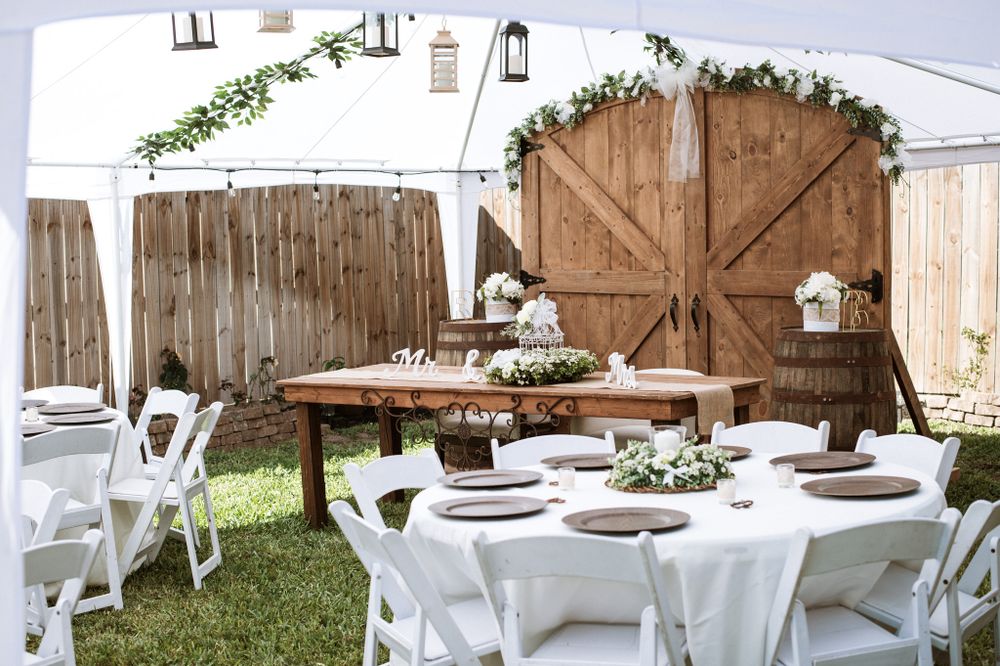 Rustic outdoor wedding reception setup with white tables and wooden barn doors decorated with greenery.