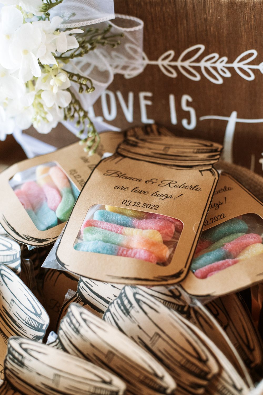 Wedding favor bags with colorful candy and 'Love Is' signage on rustic wooden surface.