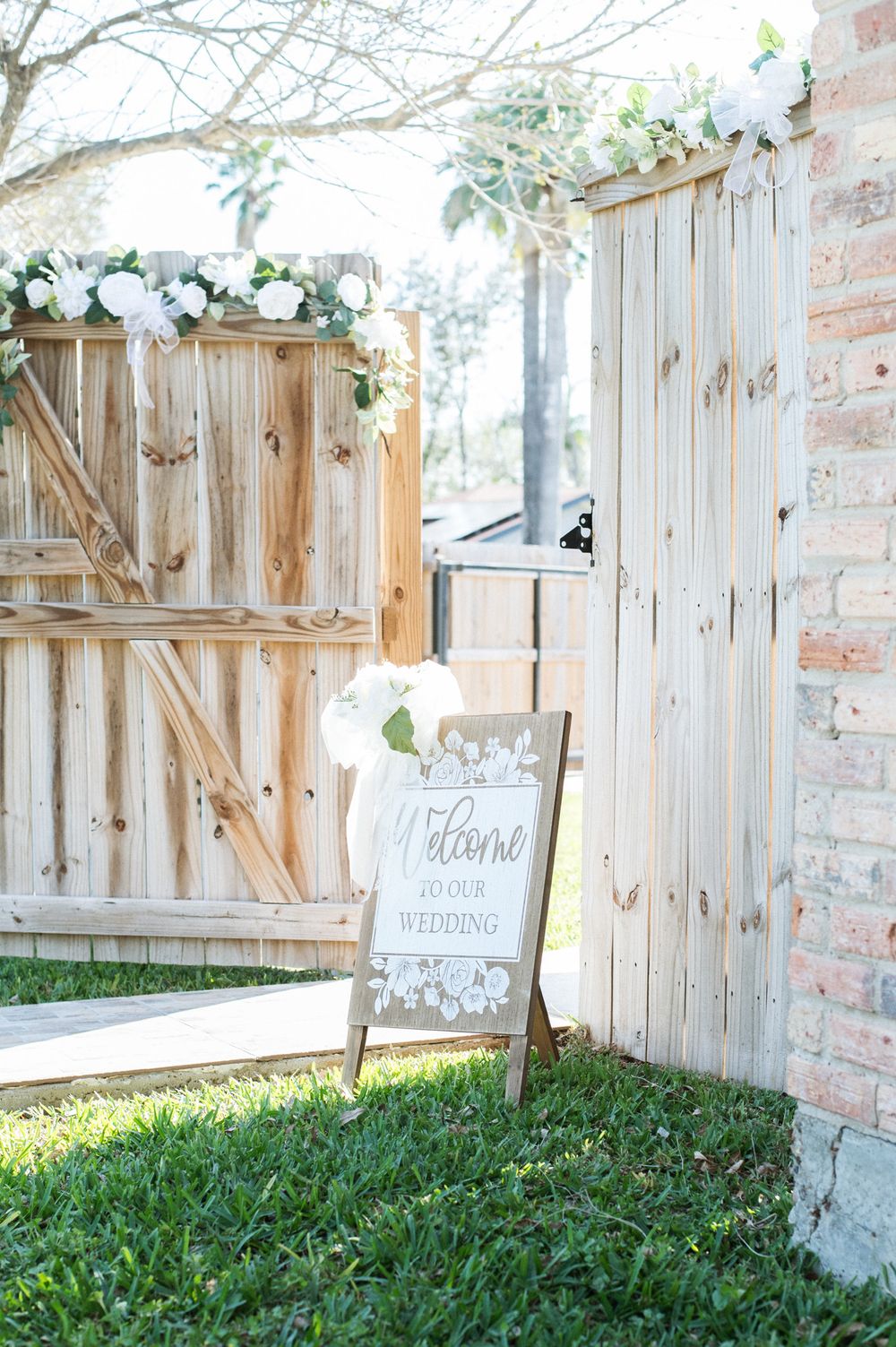 Rustic wedding welcome sign with floral decor against wooden fence backdrop.