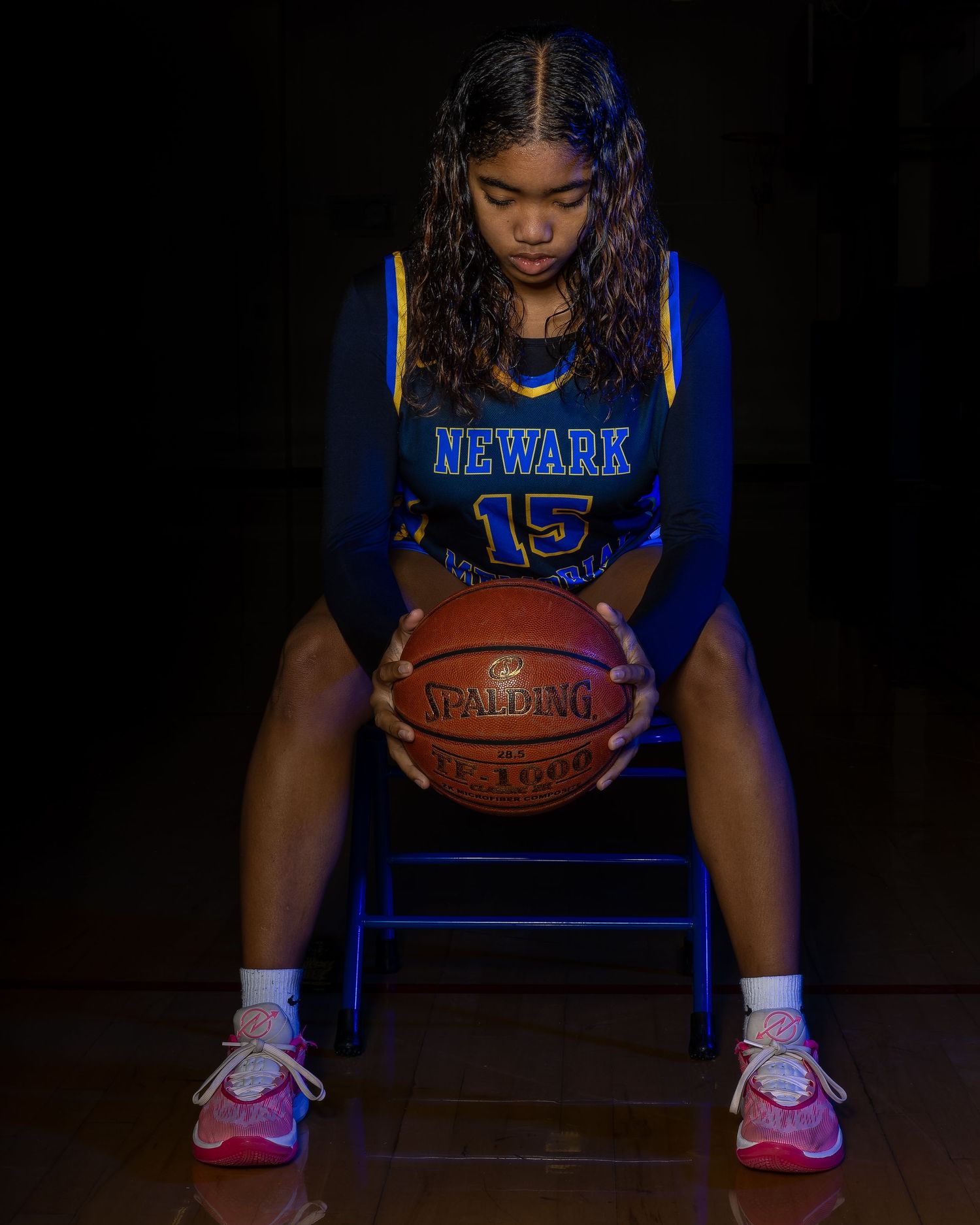 Basketball player in blue jersey number 15 poses with a basketball on a chair against dark background.