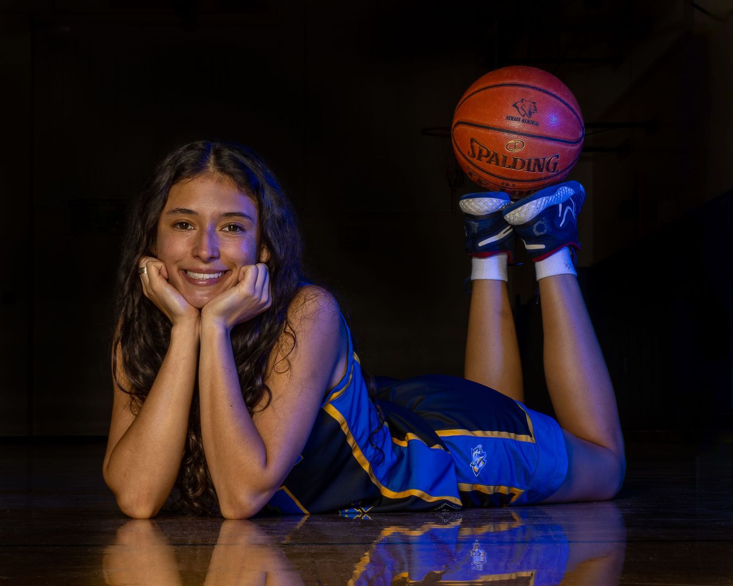 Athlete in blue uniform poses with basketball on reflective gym floor with dramatic lighting.