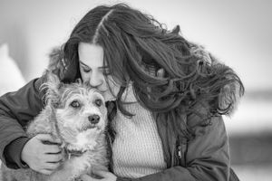 Black and white photo of a young woman kissing a small terrier dog outdoors. Pet Portrait by Xenophon Photography in Maine.